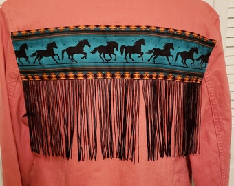 May include: A pink denim jacket with a turquoise and black patterned band with a fringe trim. The band features a row of black horses running across the band.