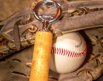 May include: A wooden baseball bat bottle opener with a silver bottle opener on top. The bottle opener is resting on a brown leather baseball glove with a white baseball inside.