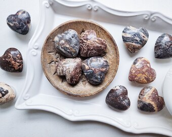 May include: A collection of nine heart-shaped polished stones with brown, black, and white swirling patterns. The stones are arranged on a white tray with a wooden bowl containing some of the stones.
