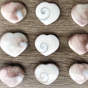 May include: Nine heart-shaped polished stones, some with a swirling pattern, others with a smooth, white surface. The stones are arranged in three rows of three on a wooden surface.