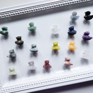May include: A collection of 16 small, hand-carved animal figurines made from various types of stone. The figurines are arranged on a white tray with a decorative border. The figurines are in various colors, including green, blue, purple, yellow, and brown.