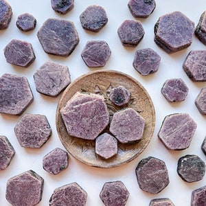 May include: A collection of purple ruby gemstone chips in various sizes and shapes. The chips are arranged on a white surface and a wooden plate.