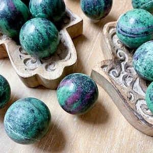 May include: A collection of green and pink zultanite spheres, also known as zultanite gemstone, in various sizes and patterns. The spheres are displayed in two wooden hand-shaped bowls.