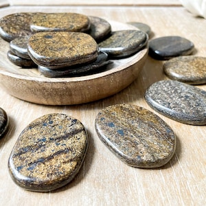 May include: A collection of smooth, oval-shaped brown and gold tiger's eye stones in a wooden bowl. The stones are polished and have a natural, striated pattern.