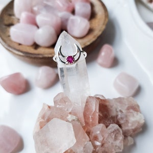 May include: A silver crescent moon ring with a pink gemstone in the center. The ring is displayed on a cluster of pink and white crystals.