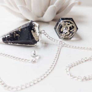 May include: Black and gold pendulum with a silver chain. The pendulum is shaped like a cone and has a hexagonal base.