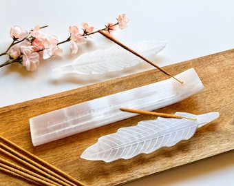 May include: White selenite crystal incense holders in the shape of feathers, on a wooden surface with incense sticks.