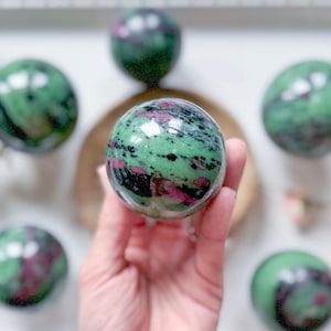 May include: A collection of polished gemstone spheres. The spheres are primarily green with dark green and pink inclusions. One sphere is held in a hand, with others arranged around it. The spheres have a glossy, reflective surface.
