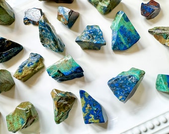 May include: A collection of rough, natural, blue and green azurite stones. The stones are various sizes and shapes, and have a mottled, textured surface.