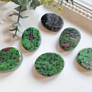 May include: Six polished oval-shaped stones with a green base color, speckled with black and red inclusions. The stones are arranged on a white surface, with a sprig of greenery in the background. The stones vary in size and shape.