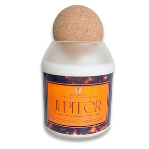 May include: A cylindrical candle with a cork stopper. The label reads "Jupiter" in orange lettering, with a galaxy design. The candle is part of the "Planetary Collection" and contains lemongrass, ginger, cardamom, patchouli, and amber.