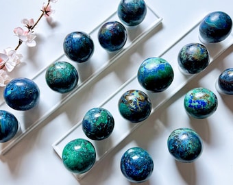 May include: A collection of 15 smooth, round, polished azurite spheres in shades of blue, green, and brown. The spheres are arranged on two white rectangular surfaces.