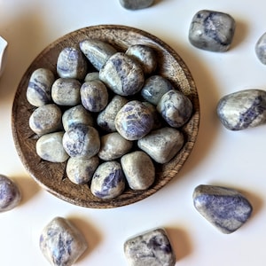 May include: A collection of tumbled blue and grey striped stones in a wooden bowl. The stones are arranged in a pile and are surrounded by other tumbled stones on a white surface.
