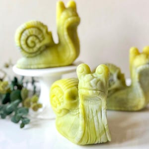 May include: Three green snail-shaped soap sculptures. The snails are made of a light green soap and have detailed carvings. The snails are arranged on a white pedestal and a white surface.