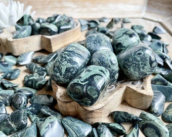 May include: A collection of green and black tumbled stones with a swirling pattern. The stones are arranged in a wooden bowl.