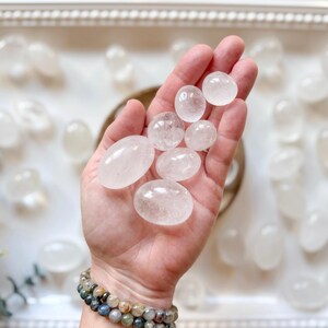 May include: A hand holding a collection of smooth, white, oval-shaped clear quartz stones. The stones are various sizes and are arranged in a pile in the palm of the hand.