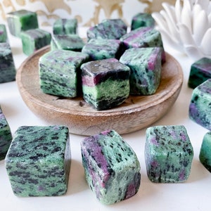 May include: A collection of green, black, and pink tumbled zultanite stones. The stones are arranged in a circle on a wooden plate.