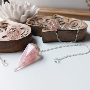 May include: Pink crystal pendulums with silver chains and accents. One pendulum is a faceted teardrop shape, and the other is a hexagonal shape. The pendulums are displayed on a white surface with wooden carved trays and a white flower.