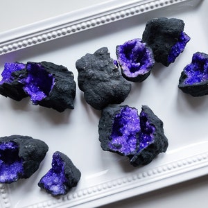 May include: A collection of dark gray geode clusters with vibrant purple crystal interiors. The geodes are arranged on a white, rectangular, decorative tray. The crystals sparkle, creating a contrast with the rough, dark exterior.