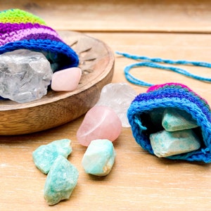 May include: Two crocheted pouches with colorful stripes, one filled with clear quartz crystals and the other with green aventurine crystals. The pouches are on a wooden surface.