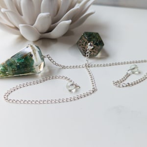 May include: A green crystal pendulum with a silver chain and a small green crystal bead. The pendulum is shaped like a cone and has a clear resin base.