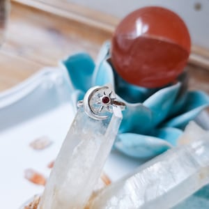 May include: A silver ring featuring a crescent moon and sun design with a small red gemstone at the center. The ring is displayed on a clear crystal, with a blue decorative flower and a red sphere in the background.