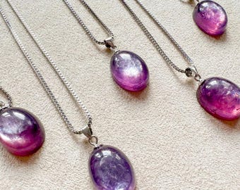 May include: Several amethyst pendant necklaces with silver chains. Each necklace features a polished, oval-shaped amethyst stone with shades of purple and pink. The necklaces are displayed on a neutral-colored surface.
