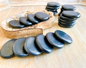 May include: A collection of smooth, oval-shaped black stones, possibly for massage or spa treatments. Some stones are stacked, others are arranged in a wooden carved dish, and some are scattered on a light-colored wooden surface.