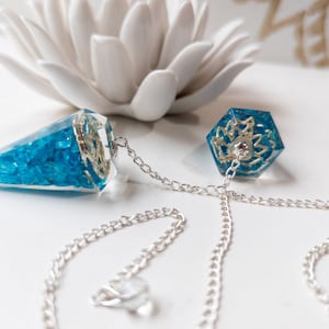 May include: A blue crystal pendulum with a silver chain. The pendulum is shaped like a cone and has a flower design on the bottom. The chain is attached to a ring at the top of the pendulum.