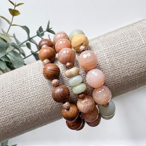 May include: A set of four bracelets featuring natural stone beads in shades of pink, peach, and green, along with brown wooden beads. The bracelets are stacked on a beige fabric surface.