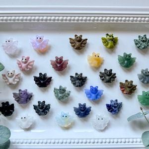 May include: A collection of 20 hand-carved crystal bats in various colors, including pink, purple, blue, green, and brown. The bats are arranged on a white surface with a decorative border.