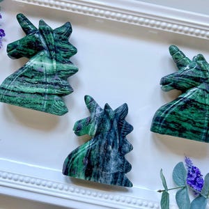 May include: Three green and black unicorn head sculptures carved from a green and black stone. The sculptures are displayed on a white surface.