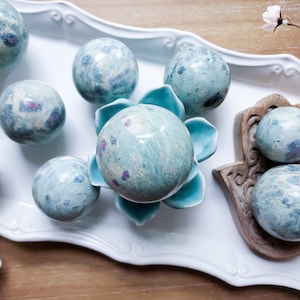 May include: A collection of polished gemstone spheres in shades of blue and green, with flecks of pink and red. The spheres are arranged on a white tray, a turquoise ceramic flower, and a wooden hand-shaped holder. Decorative and ornamental.