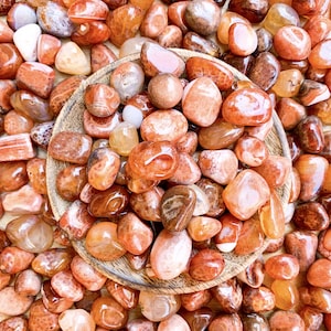May include: A collection of carnelian tumbled stones in various shades of orange and red. The stones are smooth and polished, with some showing natural banding patterns. The carnelian stones are displayed in a wooden bowl.