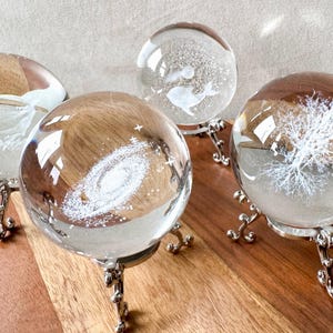 May include: Four glass paperweights on silver stands. Each paperweight features a different design: a galaxy, a tree, a fish, and a swirl pattern.