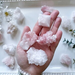 May include: A collection of pink Himalayan salt crystals of varying sizes and shapes. The crystals are clustered together and have a natural, rough texture.