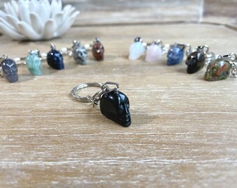 May include: A collection of various gemstone skull keychains, including black obsidian, green aventurine, blue sodalite, and pink rose quartz. The keychains are displayed on a wooden surface.