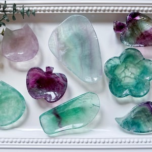 May include: Assortment of colorful fluorite crystal carvings in various shapes, including a cat, apple, leaf, and flower. The crystals display shades of purple, green, and clear, arranged on a white tray. The carvings are likely decorative items.