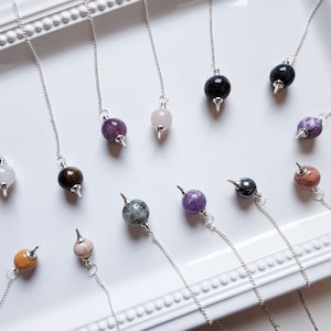 May include: A collection of 12 crystal pendulums with silver chains and various colored stones, including amethyst, rose quartz, tiger's eye, and clear quartz.