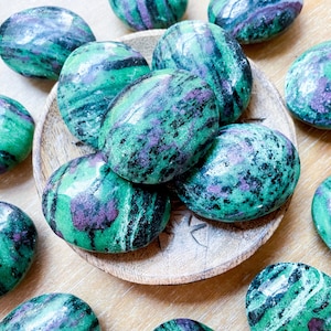 May include: A collection of green, black, and purple striped tumbled stones. The stones are smooth and polished, and they are arranged in a pile on a wooden surface.