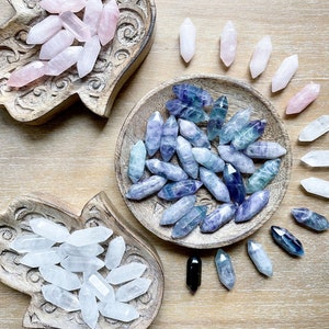May include: An overhead shot displays various crystals in wooden bowls and scattered on a wooden surface. The crystals are in shades of pink, blue, and clear, with a few black crystals. The bowls have carved designs.