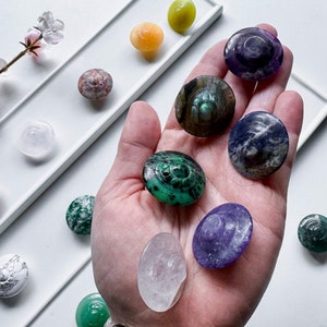 May include: A collection of various gemstone cabochons, each carved with a unique design. The cabochons are displayed on a white surface and in a hand. The gemstones include amethyst, green aventurine, labradorite, clear quartz, and more.