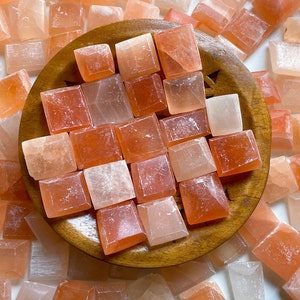 May include: A collection of small, square, pink and white selenite crystals arranged on a wooden plate. The crystals are smooth and polished, with a translucent appearance.