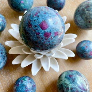 May include: A collection of eight green and pink ruby zoisite spheres of varying sizes. The spheres are arranged on a white lotus flower-shaped stand and a wooden surface.