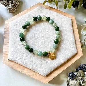 May include: A green and white beaded bracelet with a gold charm in the shape of a four-leaf clover. The bracelet is on a wooden display stand.
