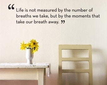 May include: A white table with a vase of yellow flowers sits next to a white chair with a checkered cushion. The wall behind the table has a quote that reads "Life is not measured by the number of breaths we take, but by the moments that take our breath away."