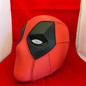 May include: A red full-head mask with a textured surface, featuring large black eye patches with white mesh lenses. The mask has defined seams across the forehead and cheek areas, creating a structured appearance with a smooth, consistent finish.