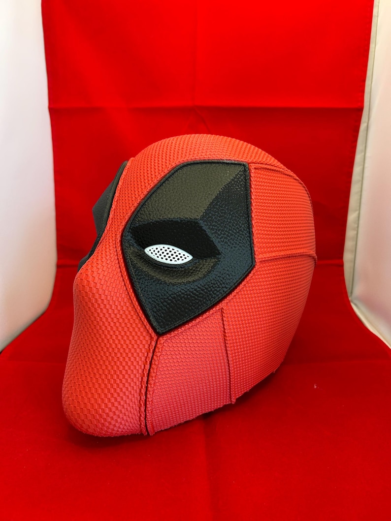 May include: A red full-head mask with a textured surface, featuring large black eye patches with white mesh lenses. The mask has defined seams across the forehead and cheek areas, creating a structured appearance with a smooth, consistent finish.