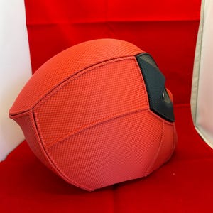 May include: A red full-head mask featuring a textured fabric surface with visible stitched seams. The mask includes a black eye section with a mesh-like material, providing a distinct contrast to the bright red fabric covering the rest of the headpiece.