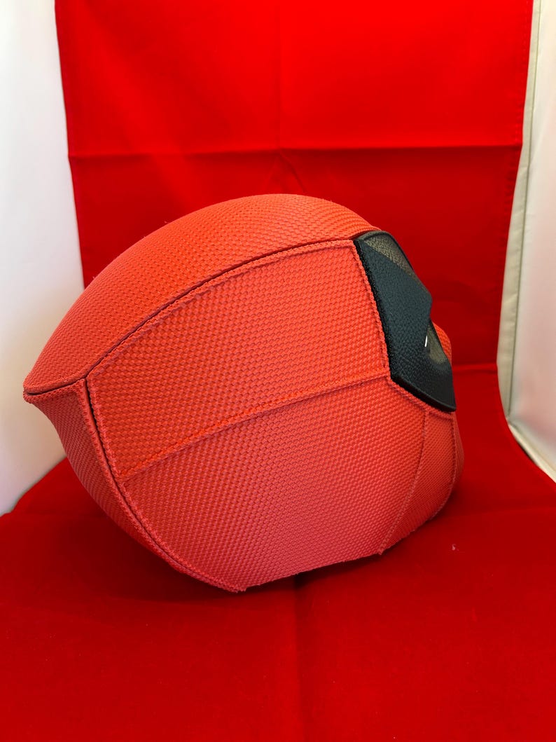 May include: A red full-head mask featuring a textured fabric surface with visible stitched seams. The mask includes a black eye section with a mesh-like material, providing a distinct contrast to the bright red fabric covering the rest of the headpiece.